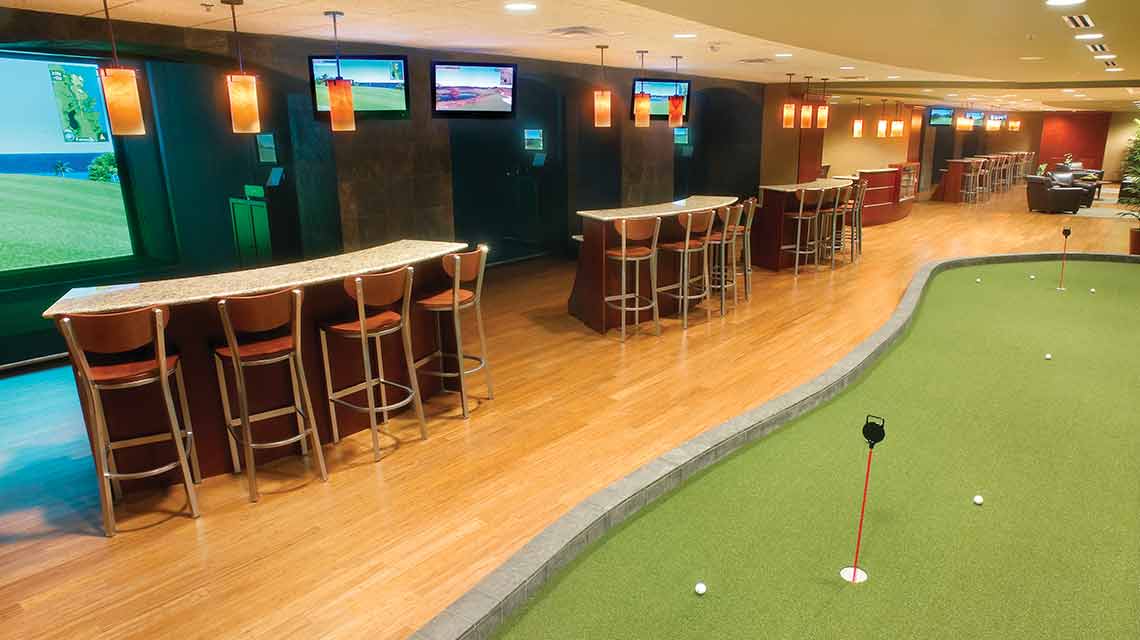 An indoor golf center featuring a putting green, a row of bar tables and bar seating and a ceiling lined with TV screens