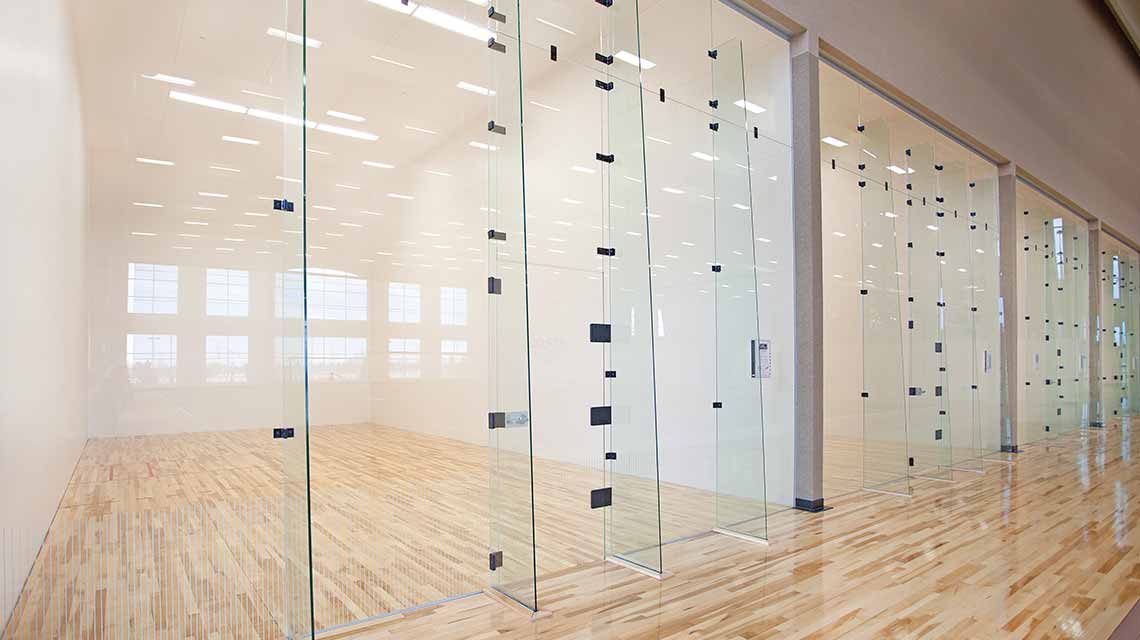 A row of four empty racquetball courts with wooden floors and glass doors for entry