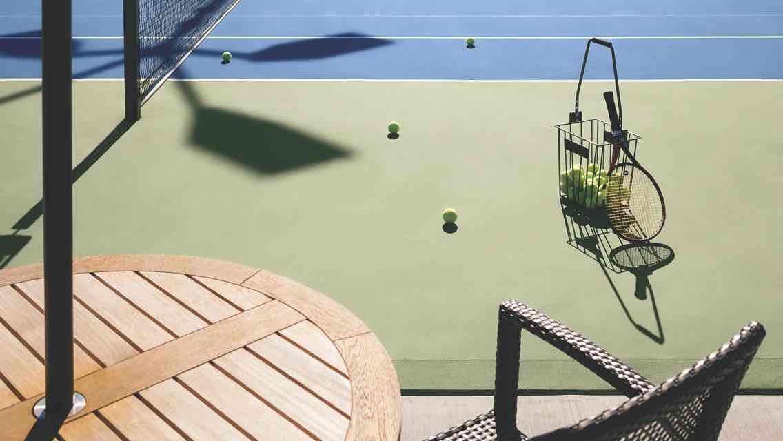 A table and chair overlook an outdoor tennis court with a basket of tennis balls and racquet standing by