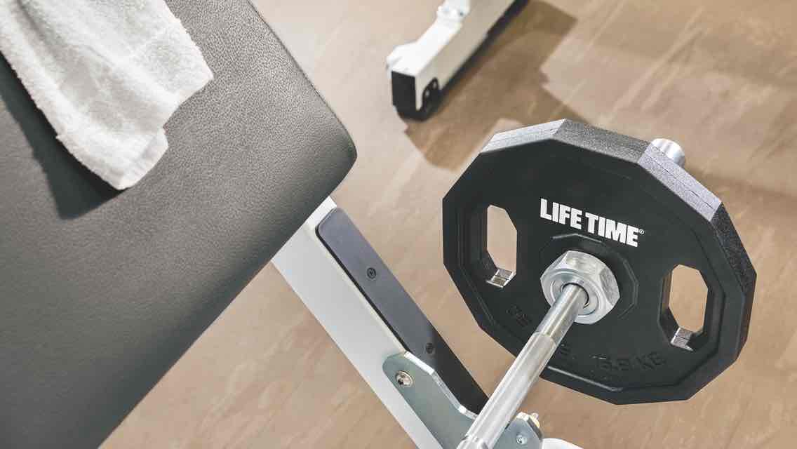 A weighted Life Time barbell and a white towel sit on a padded weight bench with a gleaming wooden fitness floor