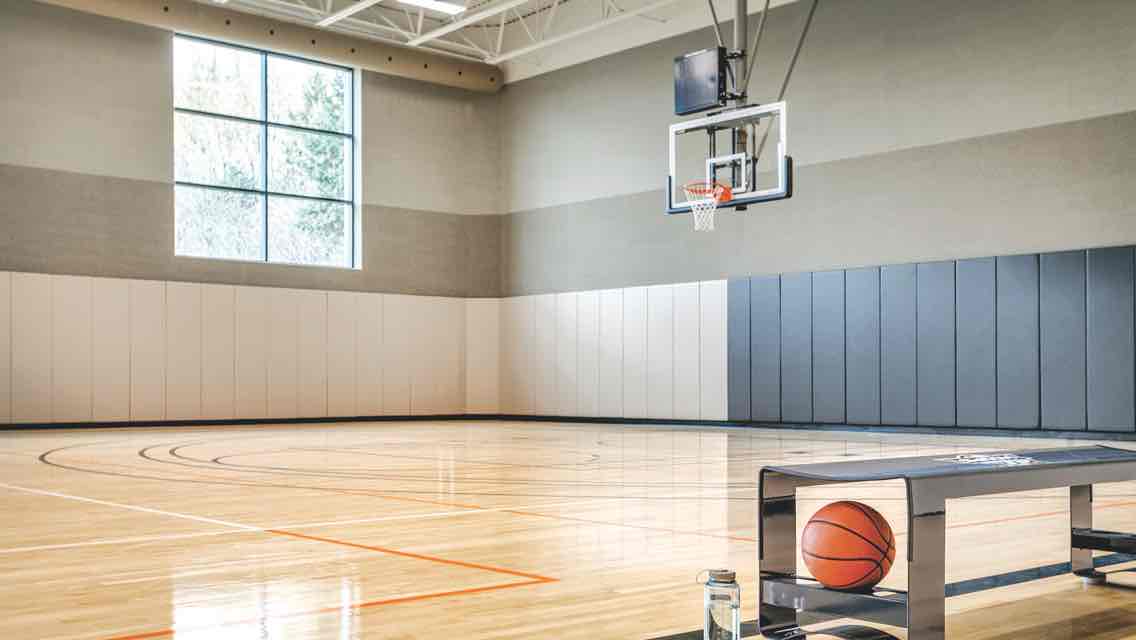 A spacious gym with large windows, a gleaming wood floor, basketball hoops and a cart filled with basketballs