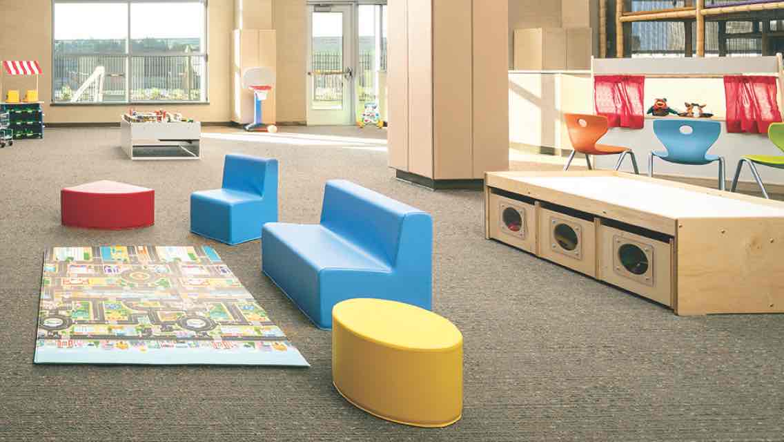 A spacious, sunlit carpeted kids play area with child seating and wooden toy containers