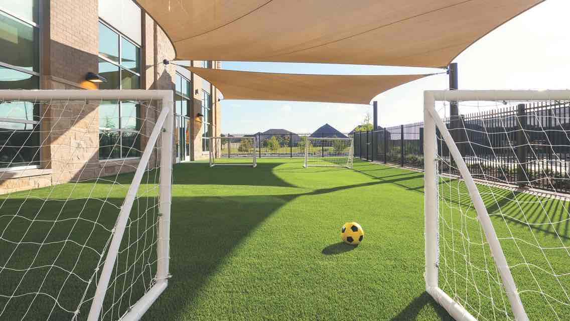 A outdoor play area with a green turf field, two soccer goals lined up and a yellow and black soccer ball