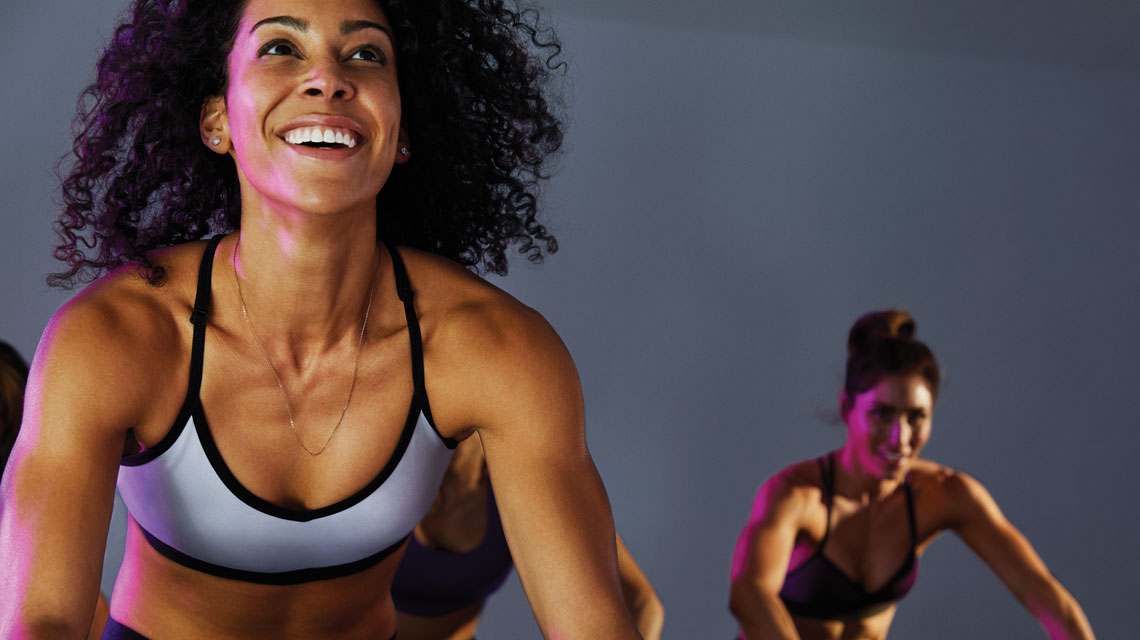 Two women in athletic clothing smiling in an indoor cycle class