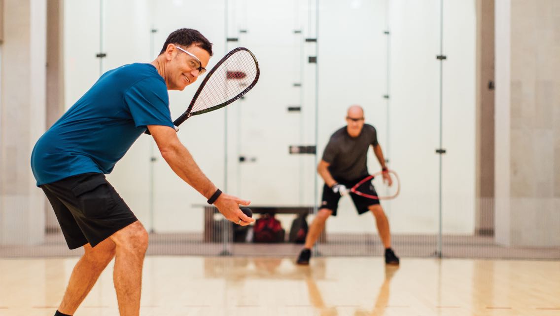 Two men in safety goggles hold racquetball rackets and face each other on the court