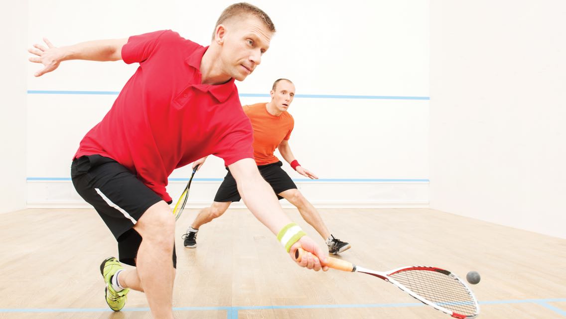 Two men holding squash racquets next to each other while one lunges toward the ball 