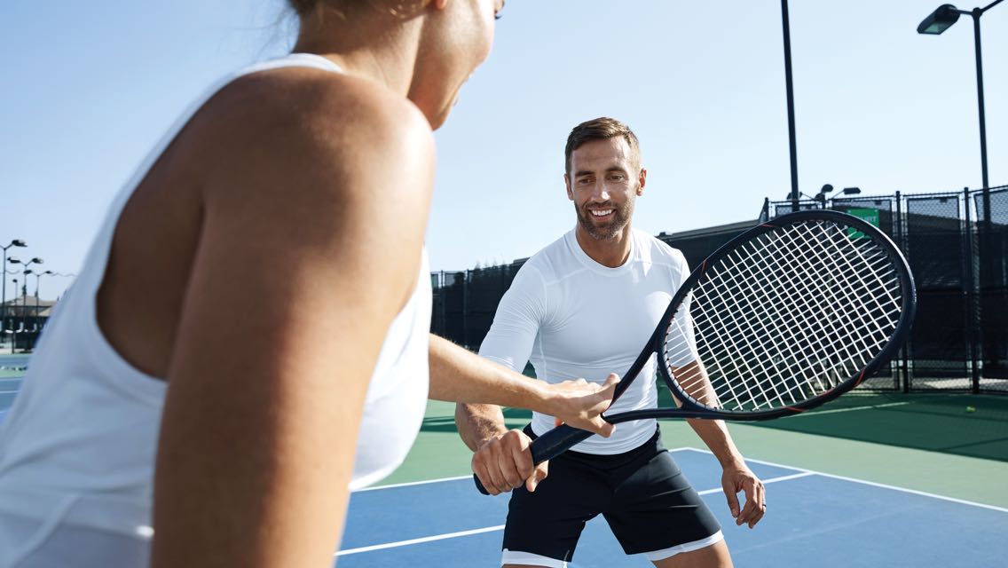 A tennis instructor hands a tennis racquet to a student while they stand on an outdoor court