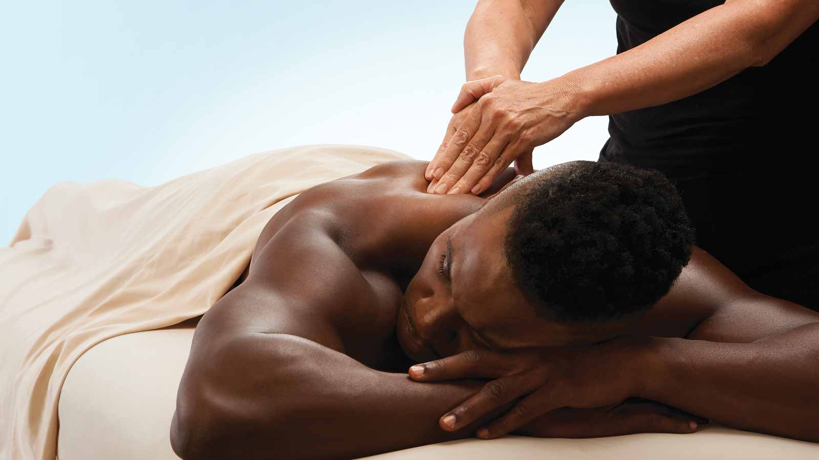 A client covered by a sheet lying face down on a massage table while a massage therapist presses their hands onto his back
