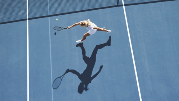 A woman in a white tennis outfit takes a one-handed backhand swing with a tennis racquet on an outdoor tennis court.