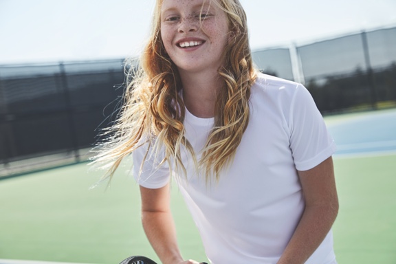 A girl with long blond hair wearing a white t shirt stands on an outdoor tennis court and smiles.