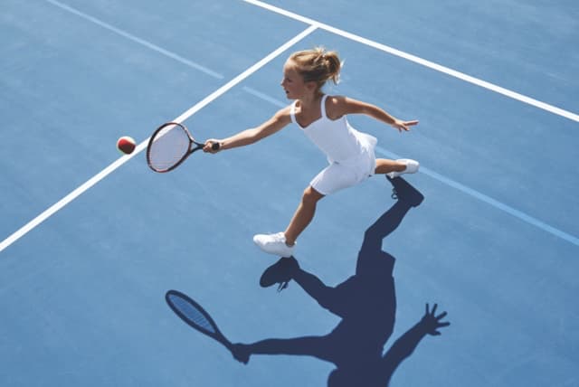 A young girl in a white tank top and white shorts takes a one-handed forehand swing with a tennis racquet at a lofted tennis ball on an outdoor tennis court.