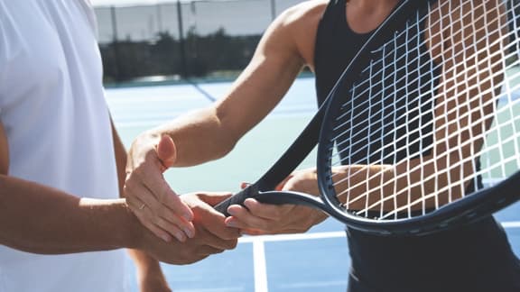 A female tennis instructor in a black tennis outfit helps a male student in a white tennis outfit perfect his grip on a tennis racquet on an outdoor tennis court.